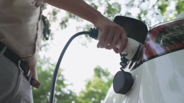 Genç adam, yaz mevsiminde açık yeşil yay yeşilliklerindeki şarj istasyonundan elektrikli araba aküsünü şarj etmek için EV şarj aleti koymuş. Yeniden şarj edilebilir elektrikli araç için sürdürülebilir enerji