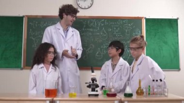 Zeki fen öğretmeni bilim teorisini çeşitli çocuklara öğretirken, mutlu ilkokul öğrencisi de mikroskop ve renkli çözeltilerle masada kimya sıvısını dinleyip öğreniyor. Pedagoji.