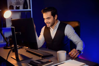 Gülümseyen zeki işadamı kahve fincanı ile uzaktan satış ekibiyle buluşuyor, gece vakti Mor Neon Light Home Office 'de bilgisayar ekranında ürün hedef pazarı hakkında araştırma yapıyor. Tahmin et..