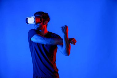 VR gözlüklü mutlu adam neon ışıklı arka planda egzersiz yapıyor ve kolunu esnetiyor. Günlük kıyafetleri olan beyaz bir insan sanal gerçeklik simülatörü teknolojisi kullanarak rahatlıyor ve egzersiz yapıyor. Sapma.