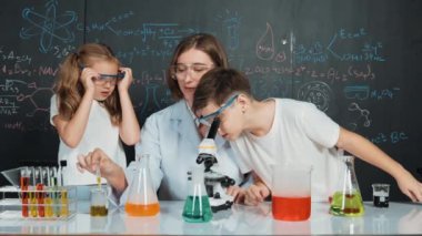 Öğretmen tavsiye verirken beyaz çocuk kimyasal sıvıyı karıştırıyor. Profesyonel eğitmen labaratuar kıyafeti giymiş, renkli solüsyonla dolu deney kabıyla sofrada çeşitli öğrencileri arıyor. Etkinlik.