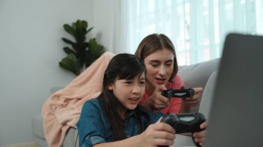 Annem dizüstü bilgisayar kullanarak kanepeye uzanırken, mutlu kız oyun oynuyor ve oturuyor. Beyaz bir ebeveyn ve çekici bir kız, elinde joystick ile birlikte vakit geçirir ve maç kazanmaya odaklanırlar. Pedagoji.