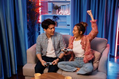 Mutlu çiftin oturduğu kanepe joysticklerle video oyunu oynuyor, meydan okuyan gülümseyen kadın, evdeki modern neon ışıklı oturma odasındaki mücadeleyi kazanmak için yumruk kaldırıyor. İnfobahn.