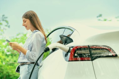 Ulusal Park veya Yaz Ormanı 'ndaki tatil gezisi sırasında EV şarj istasyonundan elektrik aküsü almak için akıllı telefon internet bankacılık uygulaması kullanan genç bir kadın. Yükselt