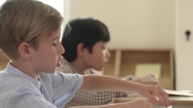 Beyaz tenli ilkokul öğrencisinin tahtada otururken bir nota bakması ve soruya cevap vermesi. Çocuklar derse katılırken genç öğretmen sınıf çalışmasını ya da sınavı açıklar. Pedagoji.