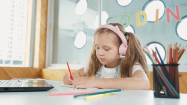 Öğretmenini dinlerken sınıfta ya da internette öğrenim gören tatlı bir kız. Öğrenci yazıları, çizimleri, bilgisayar ve renkli kalemlerle kulaklık takarak kağıt üzerinde çalışmak. Dizüstü bilgisayara odaklan. Etkinlik.