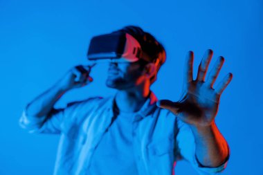 Beyaz adam ayakta durup el hareketi yapıyor ve VR gözlüğü takıyor. Gelecekçi sanal gerçeklik teknolojisi gözlüğü kullanan ve neon arka planlı sanal program gösteren gülümseyen bir genç. Sapma.