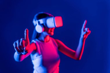 Siberpunk neon ışıklı cihazlarla çevrili zeki bir kadın metaevrene bağlı VR kulaklık takıyor, gelecekçi siber uzay teknolojisi. Sanal objeyi işaret eden bir kadın. Halüsinasyon.