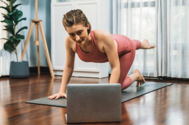 Spor kıyafetleri içinde esnek ve becerikli bir kadın evde egzersiz paspası üzerinde meditasyon pozisyonunda yoga yapıyor. Huzurlu bir zihin ve sükunetle evde yoga eğitimi..