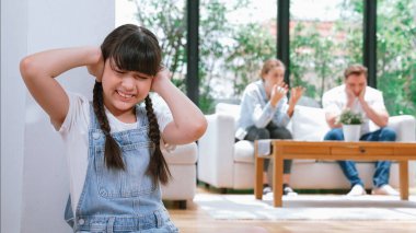 Stresli ve mutsuz genç kız köşeye kıvrılıp kulaklarını kapatarak arka planda ebeveyninin tartışmasını engelliyor. Evde aile içi şiddet ve travmatik çocukluk depresyona dönüşür. Eşzamanlama