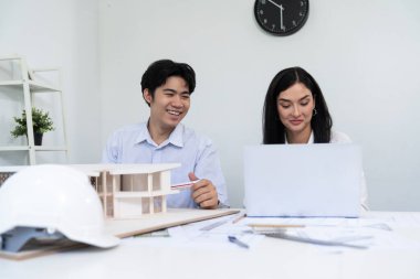 Profesyonel Asyalı erkek mimar ev modelinin uzunluğunu ölçmek için cetvel kullanırken, beyaz tenli genç meslektaşı ev modeliyle toplantı masasındaki verileri analiz etmek için dizüstü bilgisayar kullanıyor. Tertemiz..