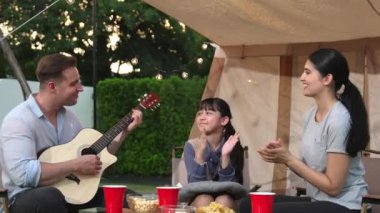 Baba gitar çalarken anne ve kız birlikte kampta oturuyorlar. Ebeveynler, genç nesil çapraz kuşak farklılıklarıyla zaman geçirmek için açık hava kamp aktivitelerini kullanıyorlar. Uyumsuzluk.