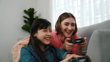 Annem dizüstü bilgisayar kullanarak kanepeye uzanırken, mutlu kız oyun oynuyor ve oturuyor. Beyaz bir ebeveyn ve çekici bir kız, elinde joystick ile birlikte vakit geçirir ve maç kazanmaya odaklanırlar. Pedagoji.
