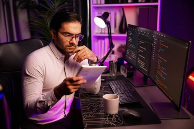 Akıllı bilişim geliştiricisi bilgisayar ekranında yazılım geliştirme kodlaması üzerinde çalışıyor ve en yeni uygulama projesinin notlarını okuyor, program güncellemesi düşünüyor neon ışıklı siber ofiste online web sitesi verileri. Tahmin et..