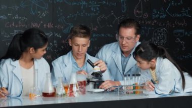 Çok kültürlü bir öğrenci mikroskop altında incelerken güzel bir kız da numuneyi inceliyor. BTMM sınıfında karatahta bilim teorisi deneyi yapan liseli çocuklara bakan öğretmen. Öğretim