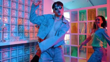 Asyalı adam gece kulübünde çeşitli neon ışıklarda dans ederken kolunu oynatıyor. Modern kulüpte çok kültürlü hippilerle dans eden şık elbiseli mutlu dansçı. Mutluluk.