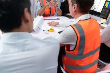 Mimari proje üzerinde birlikte çalışan çeşitli inşaat mühendisi ve müvekkil grupları, inşaat planını gözden geçirip toplantı masasında plan oluşturuyorlar. Tedbirli