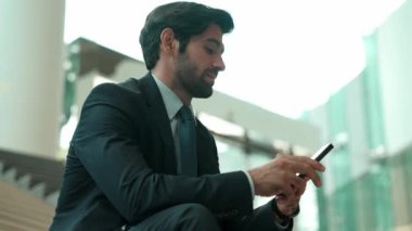 Gülümseyen yetenekli işadamı merdivenlerde otururken kameraya bakıyor. Genç profesyonel proje yöneticisi arka planda bulanık cep telefonunu tutarken kameraya gülümsüyor. Sevinçli..