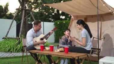 Baba gitar çalarken anne ve kız birlikte kampta oturuyorlar. Ebeveynler, genç nesil çapraz kuşak farklılıklarıyla zaman geçirmek için açık hava kamp aktivitelerini kullanıyorlar. Uyumsuzluk.