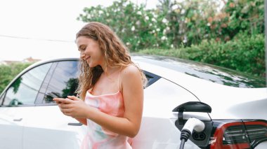 Genç bir kadın doğadaki EV şarj istasyonunda elektrik için akıllı telefon kullanıyor. Modern çevre ve EV araçlı sürdürülebilir otomobil taşıma yaşam tarzı. Eşzamanlama