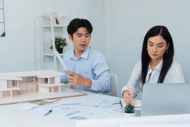 Profesyonel Asyalı erkek mimar ev modelinin uzunluğunu ölçmek için cetvel kullanırken, beyaz tenli genç meslektaşı ev modeliyle toplantı masasındaki verileri analiz etmek için dizüstü bilgisayar kullanıyor. Tertemiz..