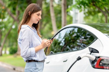Genç kadın EV şarj istasyonundan EV elektrikli araç aküsünü şarj ediyor ve şehir parkındaki elektriği ödemek için akıllı telefon internet bankacılığını kullanıyor. EV arabalı çevre dostu bir araç. Yükselt