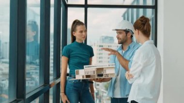 Proje yöneticisi ev modelini elinde tutarken, mimar mühendis takımı da ev tasarımı veya bina inşaatı hakkında fikir alışverişinde bulunuyor. Şehrin penceresinin yanında iç mimar duruyor. İzleme