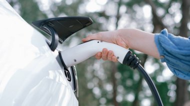 Kapalı alan kadını, doğal yeşil parktaki otoparkta EV elektrikli araba aküsünü şarj ediyor. Sürdürülebilir ve çevre dostu seyahat için şarj edilebilir araç için temiz enerji teknolojisi. Kalıcı