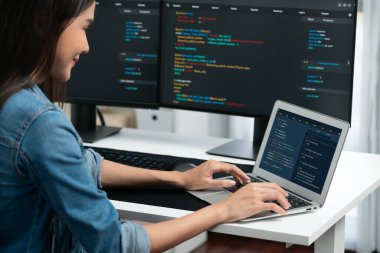 IT geliştiricisinde gülümseyen genç Asyalı dizüstü bilgisayarında kodlama programı verileriyle kotlu tişört giyerek çevrimiçi bilgi yazıyor. Güvenlik analizi iki ekran ile çevrili. Taktik.