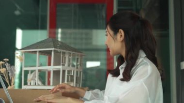 Genç ve güzel mimar mühendisin portresi çatı malzemelerini kontrol ediyor ve ofisteki masaya yerleştirilmiş proje planı ve ev modeliyle dizüstü bilgisayar kullanıyor. Yaratıcı iş tasarımı konsepti. Yönlendirici.