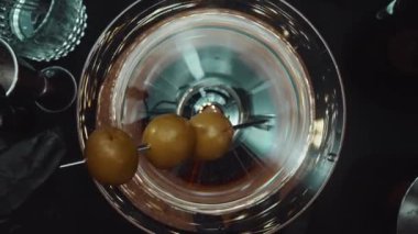 Üstten aşağıya yetenekli barmen eli zeytinli martini hazırlıyor. El yapımı şeffaf kokteyl, zeytinli, hareketli ve siyah arka planlı. Makrografi. Geliştirilebilir.