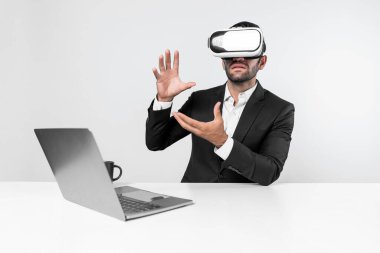 Profesyonel proje yöneticisi dizüstü bilgisayar başında otururken VR gözlüğü kullanıyor. Görsel gerçeklik kulaklığı takan yetenekli bir iş adamı metaevren ya da görsel dünyayı birbirine bağlıyor. Yenilik. Sapma.