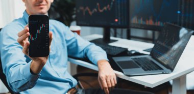 Akıllı telefonda dinamik borsa grafiği gösteren başarılı genç tüccar, bilgisayarın bulanık arka planında gerçek zamanlı döviz kuru veri tabanı sunuyor. Toplayıcı.