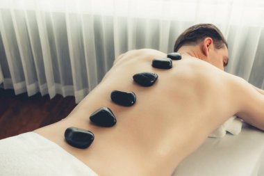 Lüks tatil köyündeki kaplıcada sıcak taş masajı. Gün ışığı dinginliği ortamı, mutlu erkek müşteri bazalt taşı masajının keyfini çıkarıyor. Rahatlatıcı bir sıcaklıkla vücudun üzerinde süzülüyor. Sessiz.