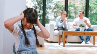 Stresli ve mutsuz genç kız köşeye kıvrılıp kulaklarını kapatarak arka planda ebeveyninin tartışmasını engelliyor. Evde aile içi şiddet ve travmatik çocukluk depresyona dönüşür. Eşzamanlama