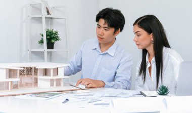 Profesyonel Asyalı erkek mimar ev modelinin uzunluğunu ölçmek için cetvel kullanırken, beyaz tenli genç meslektaşı ev modeliyle toplantı masasındaki verileri analiz etmek için dizüstü bilgisayar kullanıyor. Tertemiz..