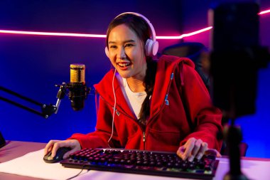 İnternet üzerinden kulaklık takıp gülümseyen Asyalı güzel kız flamasının sunucu kanalı izleyicilerle internet üzerinden konuşma yapıyor. Esport yetenekli takım oyuncuları neon mavi odada. Taktik.