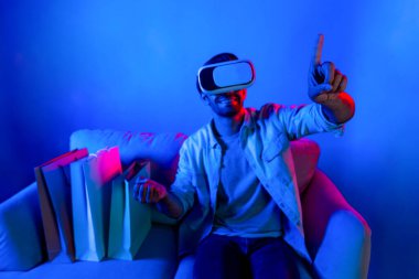 Video kameralı beyaz bir adam kanepeye oturmuş alışveriş torbalarıyla işaret ediyor. VR teknolojisi ve evde sanal gözlük kullanarak online alışveriş yapan kişi. Sapma.