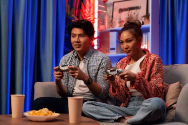 Neşeli, gülümseyen bir çiftin koltuğunda oturup oyun oynuyorlar. Oyun konsolu, hazır aperatifler ve içecekler, modern neon ışıklı, rahat oturma odasında eğlenceli bir rekabet seviyesinde. İnfobahn.