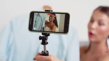 Genç sosyal medyum kadın moda videosu yapıyor. Blogcu kameraya selfie çubuğu ve ışık halkası kullanarak gülümserken, aynı zamanda ikna edici çevrimiçi kıyafetlerin izleyicilere veya takipçilere vlog satmasını sağlıyor. Düzenle