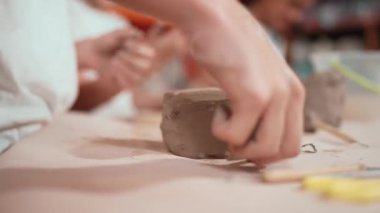 Lisedeki çocuğun el resmi. Kil oyma aleti kullanıyor. Çeşitli çocuklar çömlekçilik atölyesinde oyun oynarken ya da modellik yaparken etkinlik gösteriyor. Geçmişi bulanıklaştırıyor. Öğretim