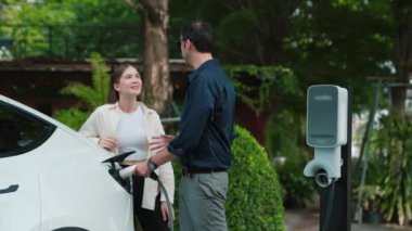 Yavaş çekim çift, ilkbaharda açık yeşil şehir kafesindeki şarj istasyonundan elektrikli araba aküsünü şarj ediyor. Sürdürülebilir çevre dostu seyahat yaşam tarzı için şarj edilebilir EV araba