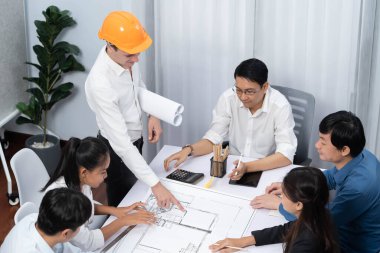 Mimari proje üzerinde birlikte çalışan çeşitli inşaat mühendisi ve müvekkil grupları, inşaat planını gözden geçirip toplantı masasında plan oluşturuyorlar. Tedbirli