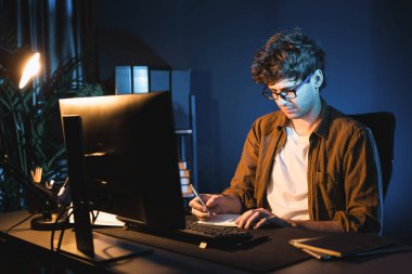 Genç işadamı, Neon Light Modern Ofis 'teki yeni proje surround çalışma masası için kağıt üzerinde tasarım düşünüyor. E-posta ile hediye göndermeden önce not defterine yeni bir iş düşünüyorum. Toplayıcı.