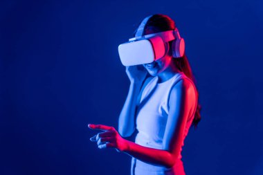 Siberpunk neon ışıklı cihazlarla çevrili zeki bir kadın metaevrene bağlı VR kulaklık takıyor, gelecekçi siber uzay teknolojisi. Sanal objeyi işaret eden bir kadın. Halüsinasyon.