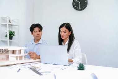 Professional male asian architect using ruler to measure house model length while young beautiful caucasian colleague using laptop to analyzed data on meeting table with house model. Immaculate. clipart