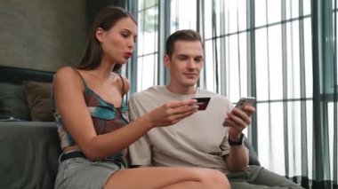 Genç çift, kredi kartıyla ödemek için internet üzerinden ödeme uygulaması ve dijital cüzdan kullanarak yatak odasında oturuyor. Mobil internet üzerinden alışveriş ve modern alımlar. Düzenle