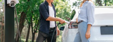 Odaklanmış EV arabanın pili, tatilde ulusal park ve yeşillik ormanlarında çevre dostu elektrikli arabalarla seyahat eden sevgili çiftin bulanık arka planına şarj oluyor. Yükselt
