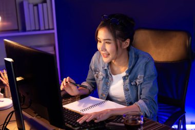 Smiling young beautiful asian creative looking on pc of profit of sales target report and writing on notebook for memo with planning invest new strategy market project at neon light night. Stratagem. clipart