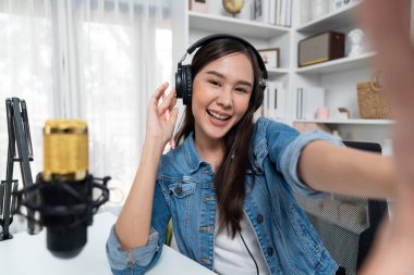 Sunucu kanalı genç ve güzel Asyalı kız akıllı telefonuyla selfie çekiyor. Modern stüdyo ofis konseptinde kulaklık takıyor. Sahne arkası yayıncısı sosyal medyada canlı yayın yapıyor. Taktik.